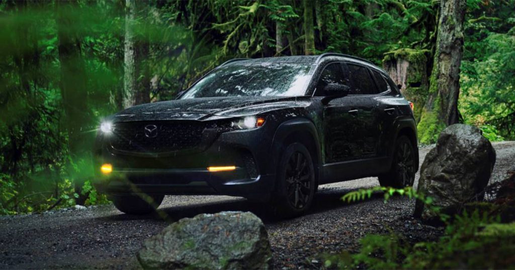 The new 2026 Mazda CX-50 drives through a shady forest with its headlights beaming to light the way. 